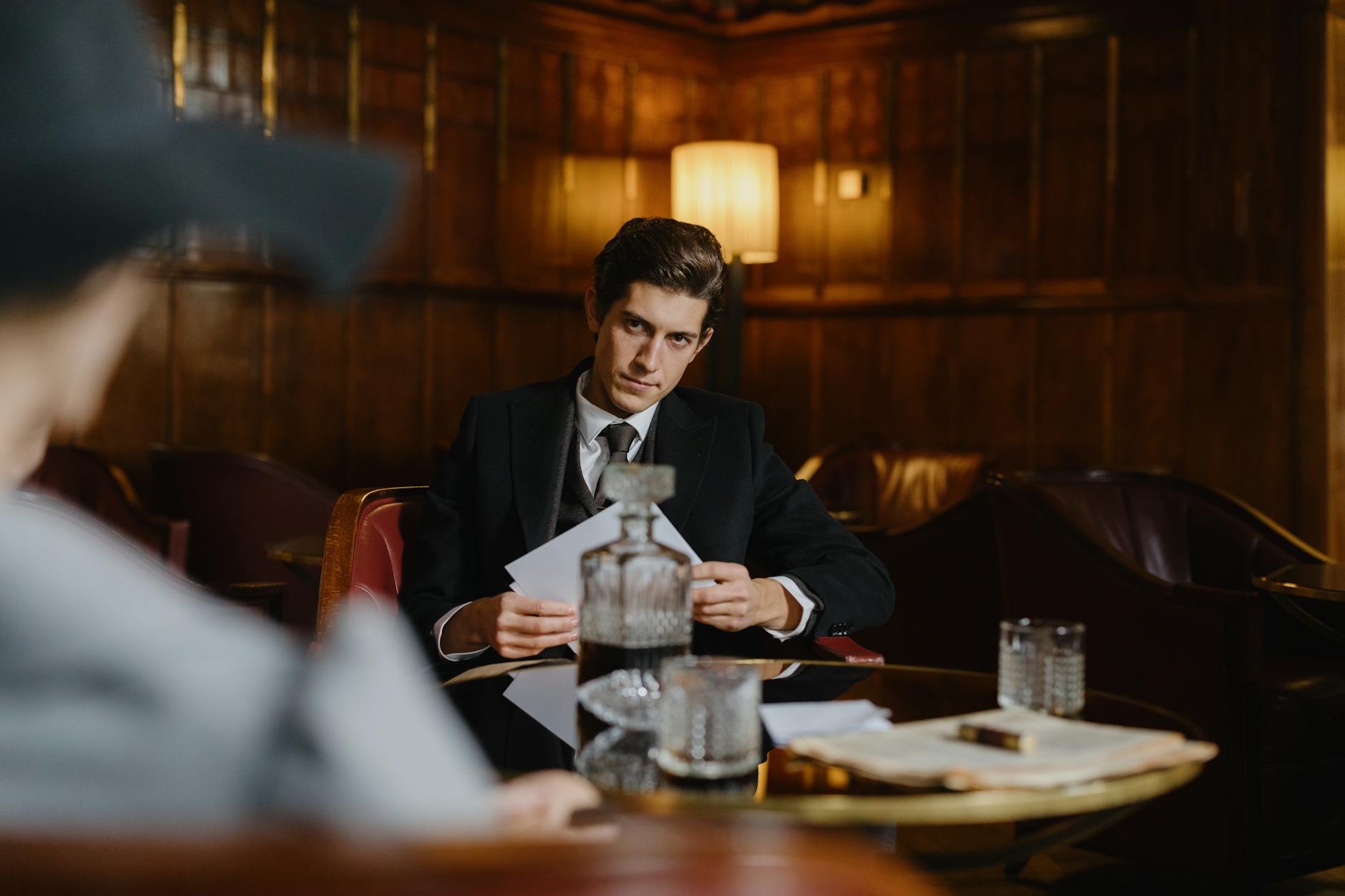 a man in black suit sitting near the table while holding a paper